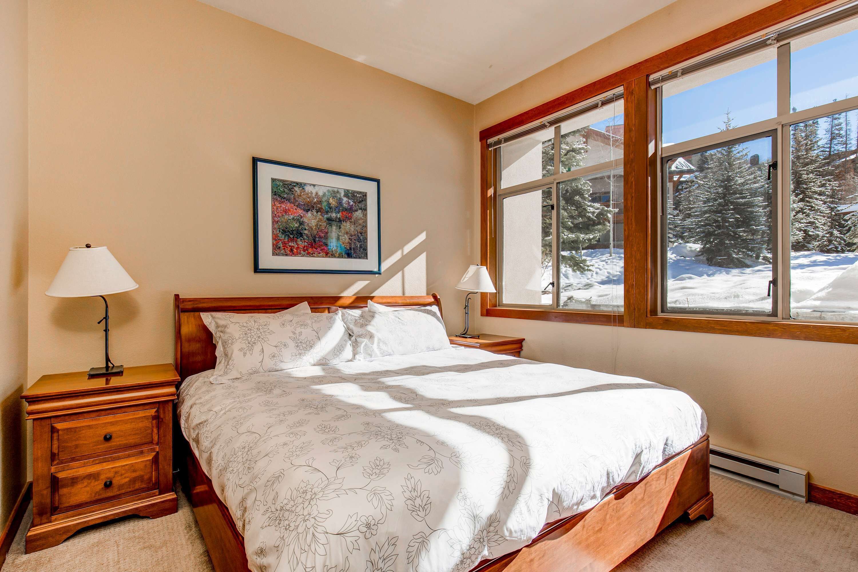 A bedroom with a bed, two bedside tables, a window with a view of snow-covered trees, and a framed picture on the wall.