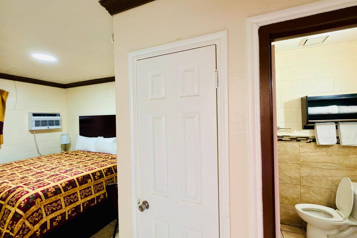 Una habitación de hotel con cama, puerta, baño con inodoro y lavamanos, y un perchero para toallas.