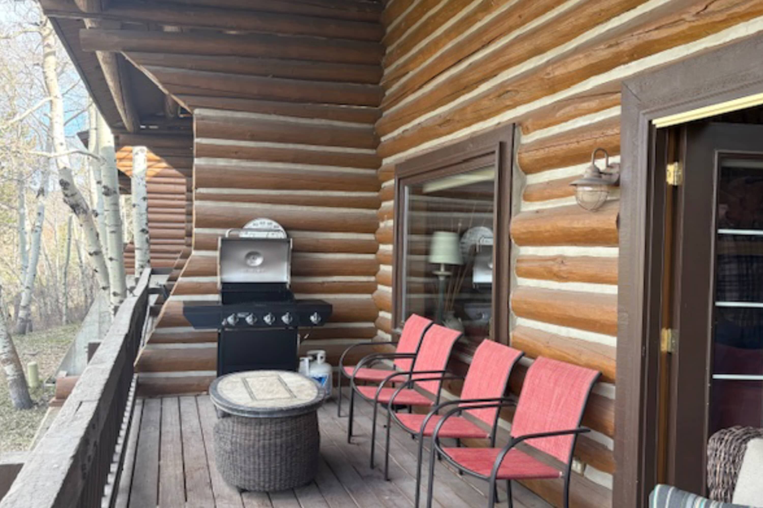 A log cabin with a deck, outdoor grill, wicker table, and red chairs.