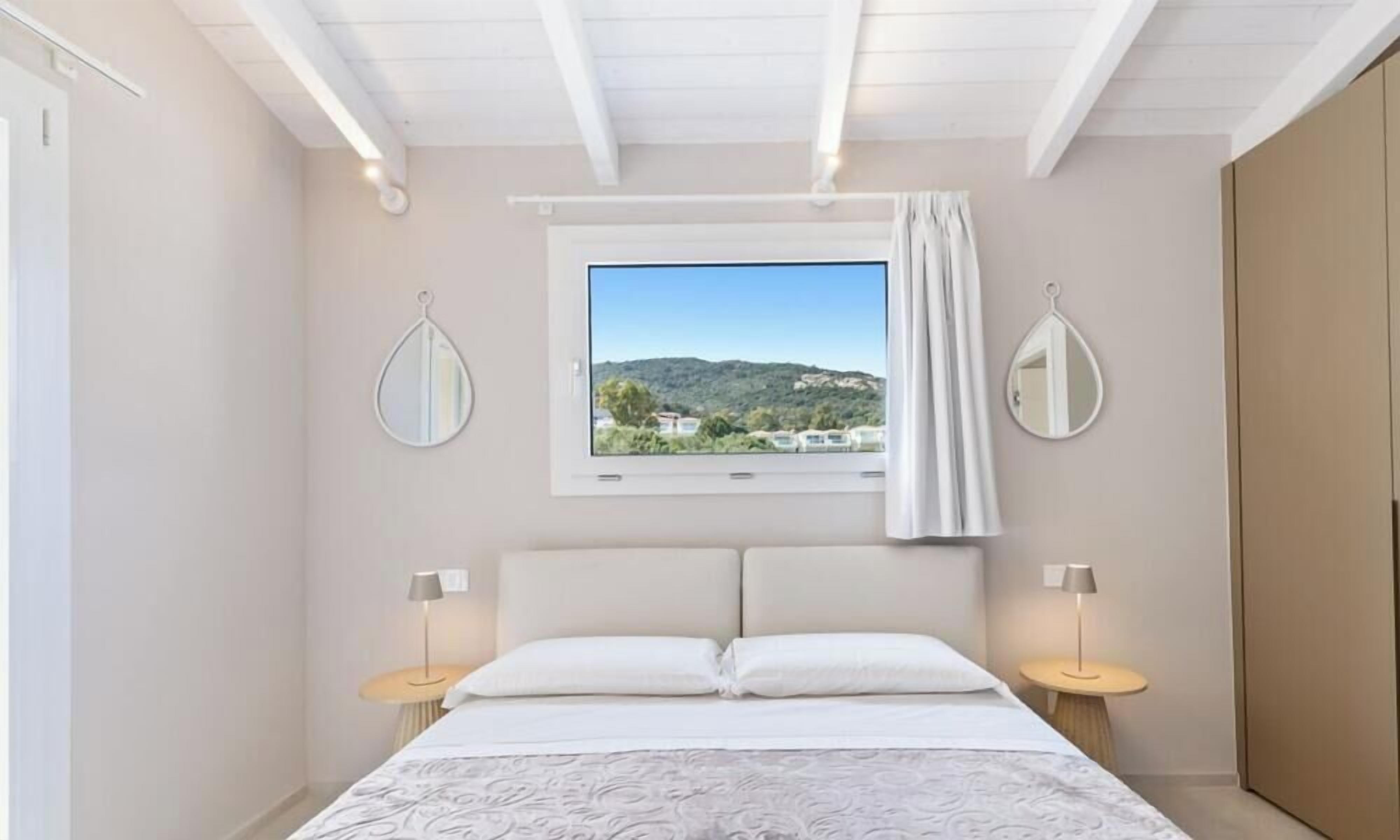 A bedroom with a bed, two bedside tables, a window with a view, and two wall-mounted mirrors.