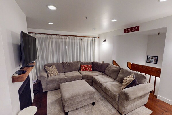 A living room with a sectional sofa, a flat-screen TV, a wooden shelf, and a staircase. - Dog-friendly Old Town Empire Condo near resort with  ski storage (Park City)