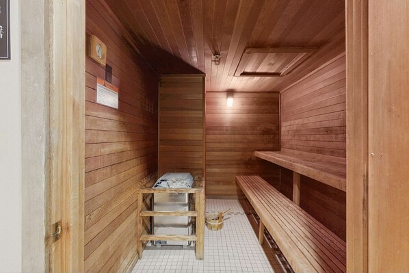 Una sala de sauna con paredes de madera, bancos, un estante metálico y suelo de baldosas.