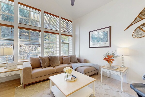 A modern living room with a beige sofa, a coffee table with a vase of flowers, a side table with a lamp, and large windows with blinds. - Relaxing mountain-view condo with shared hot tub, pool & more-walk to ski lifts (Solitude)