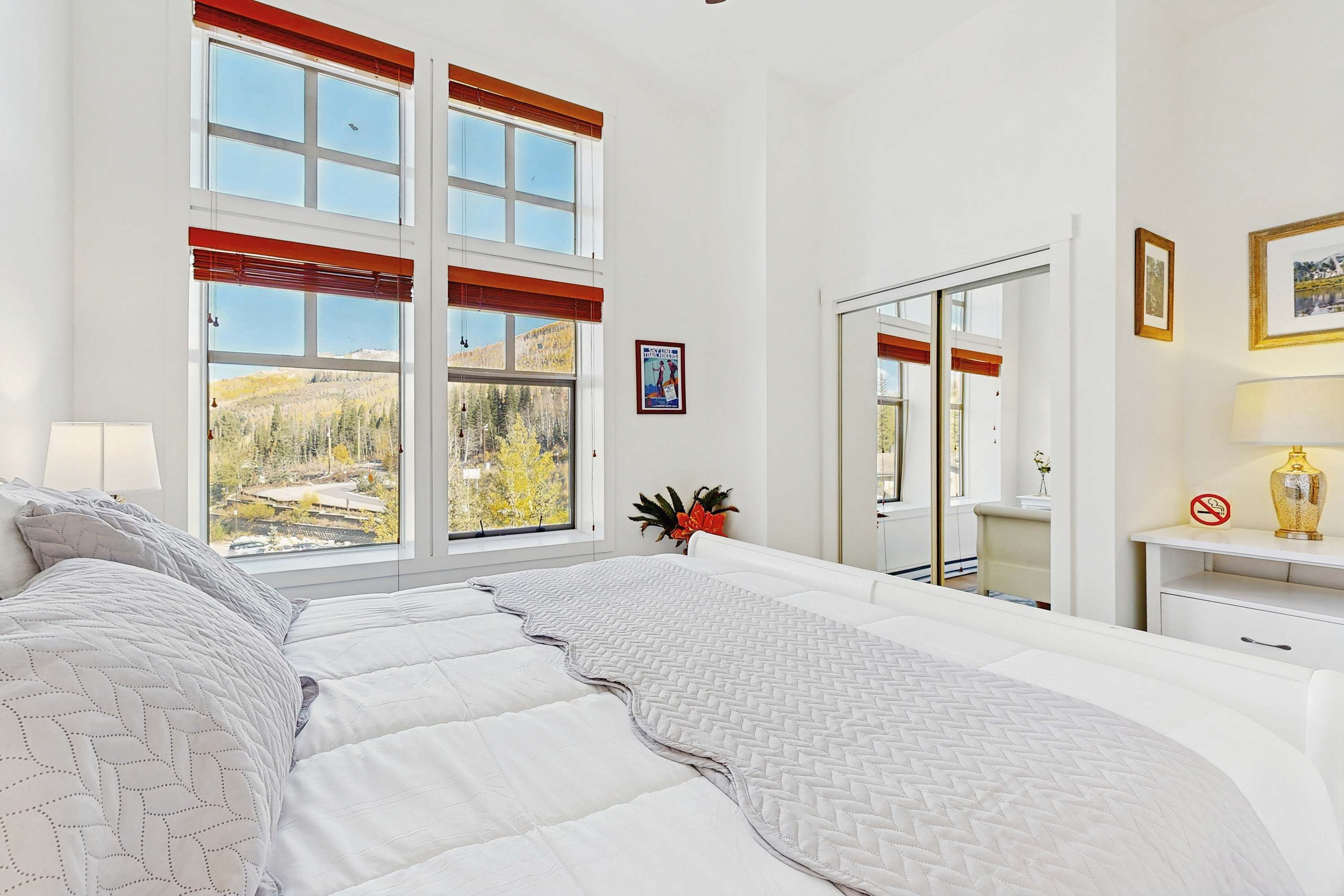 A bedroom with a large window, a bed, a bedside table, and a mirror.