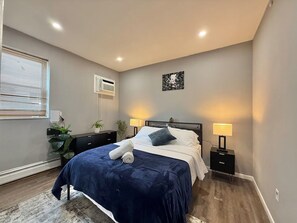 Une chambre à coucher avec un lit, des lampes de chevet, un climatiseur fixé au mur, une fenêtre avec des stores et une petite plante.