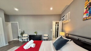 A hotel room with a bed, a red throw blanket, a white towel, a desk, two chairs, a small plant, and a shower area. - Modern apartment close to NYC (North Bergen)