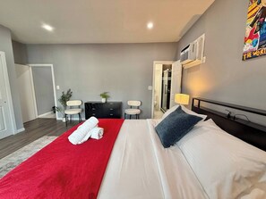 Une chambre d’hôtel avec un lit, un plaid rouge, une serviette blanche, un bureau, deux chaises, une petite plante et un espace douche.
