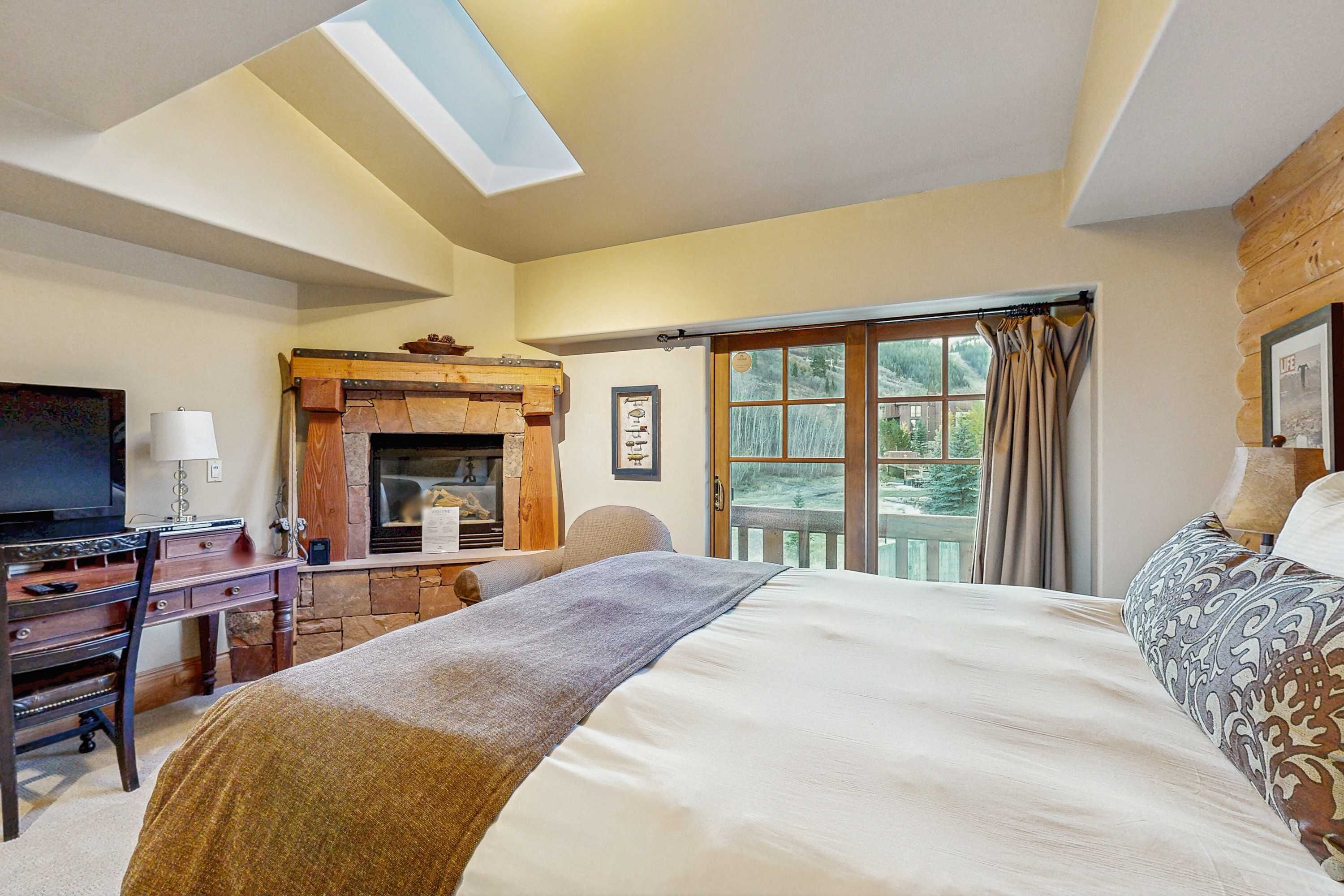 A bedroom with a bed, a desk, a television, a fireplace, and a large window with curtains.