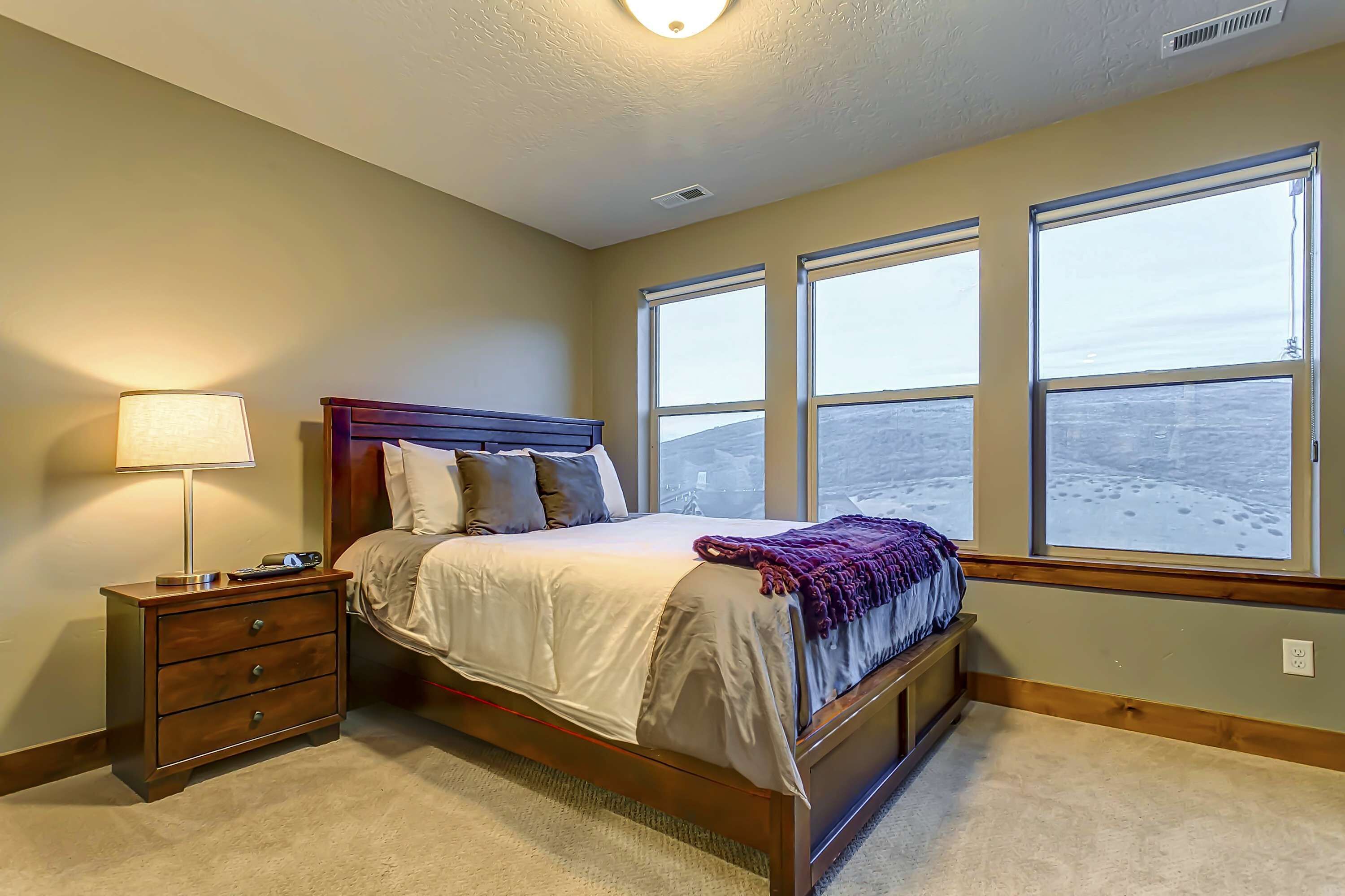 A bedroom with a bed, nightstand, lamp, and large windows.