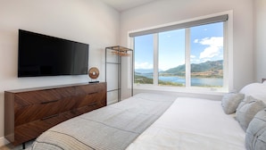 A bedroom with a large bed, a TV mounted on the wall, a wooden dresser, and a window with a view of a lake and mountains. - Townhouse in Hideout with foosball table (Hideout)