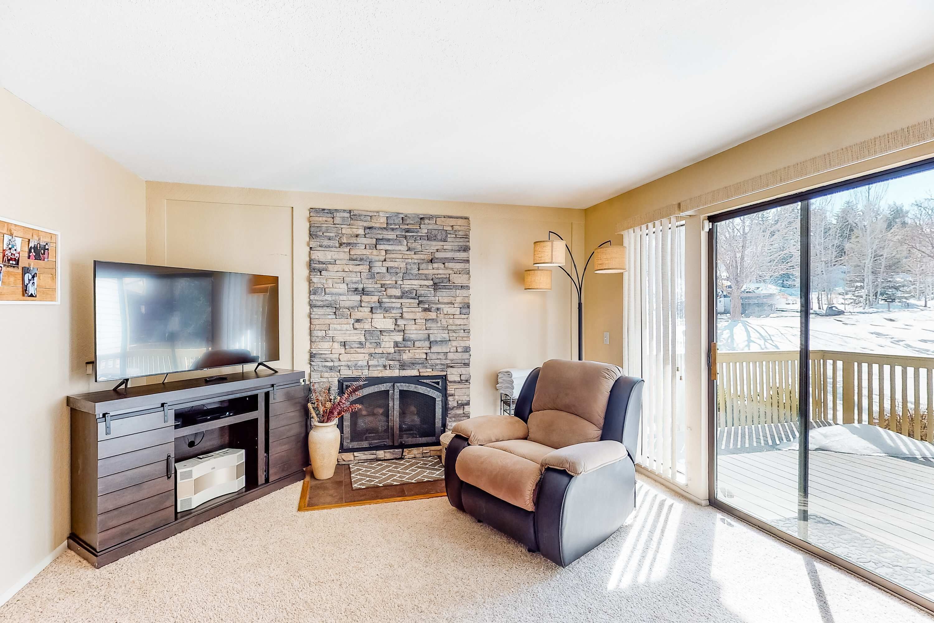 A living room with a stone fireplace, a television, a recliner, and a view of a snowy deck.