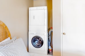 A compact, white wall-mounted washer and dryer unit in a small laundry room with a wicker headboard and striped bedding. - Three-Story, Dog-Friendly Townhouse with  Free WiFi, Gas Fireplaces, & washer & dryer (Park City)