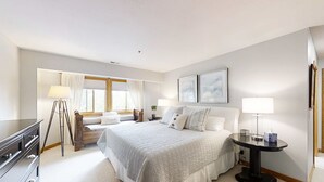 Une chambre à coucher comprenant un lit, des tables de chevet, une commode, une lampe, une fenêtre avec des rideaux et des œuvres d’art encadrées accrochées au mur.
