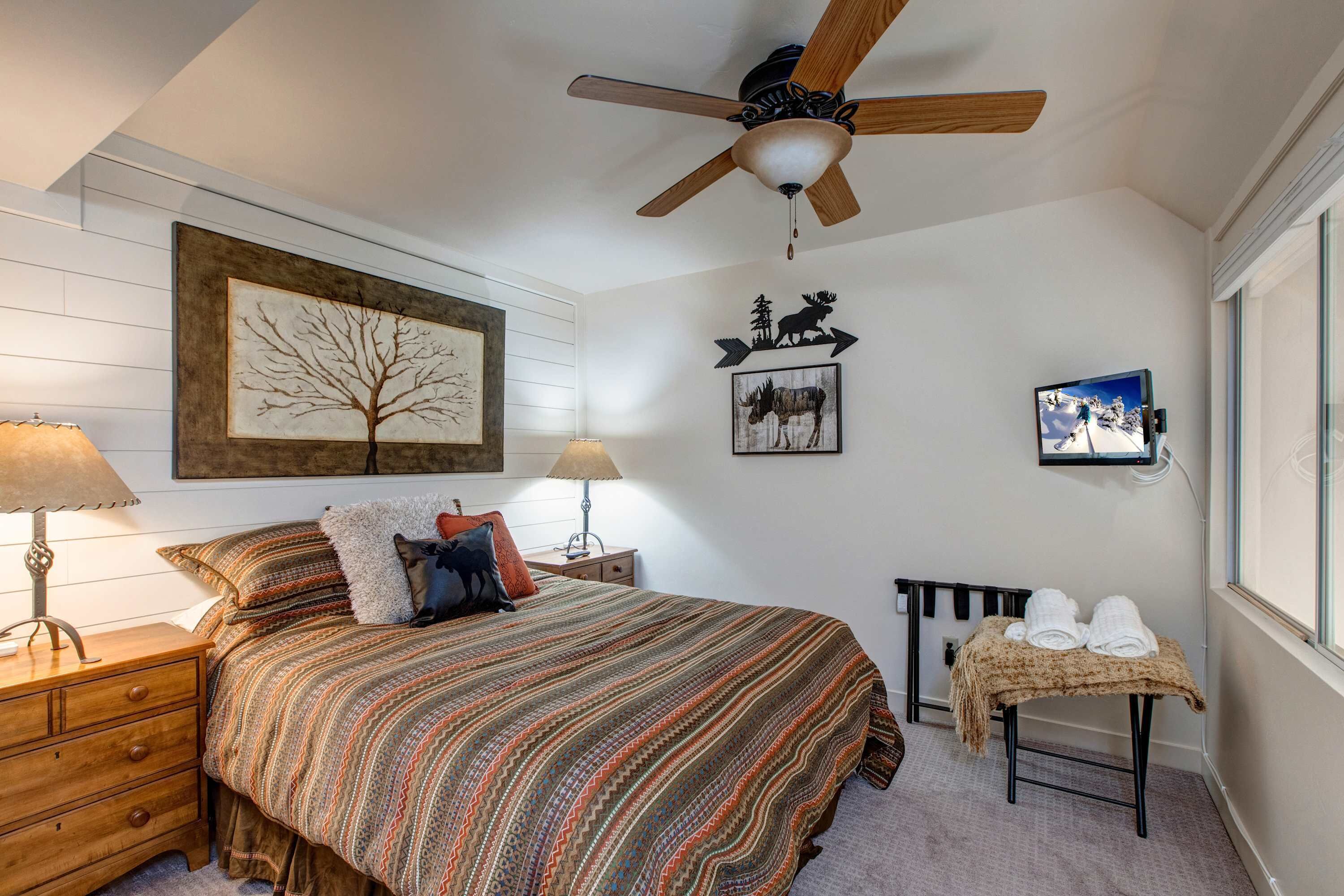 A bedroom with a bed, a ceiling fan, a nightstand, a lamp, a wall-mounted TV, and a window.