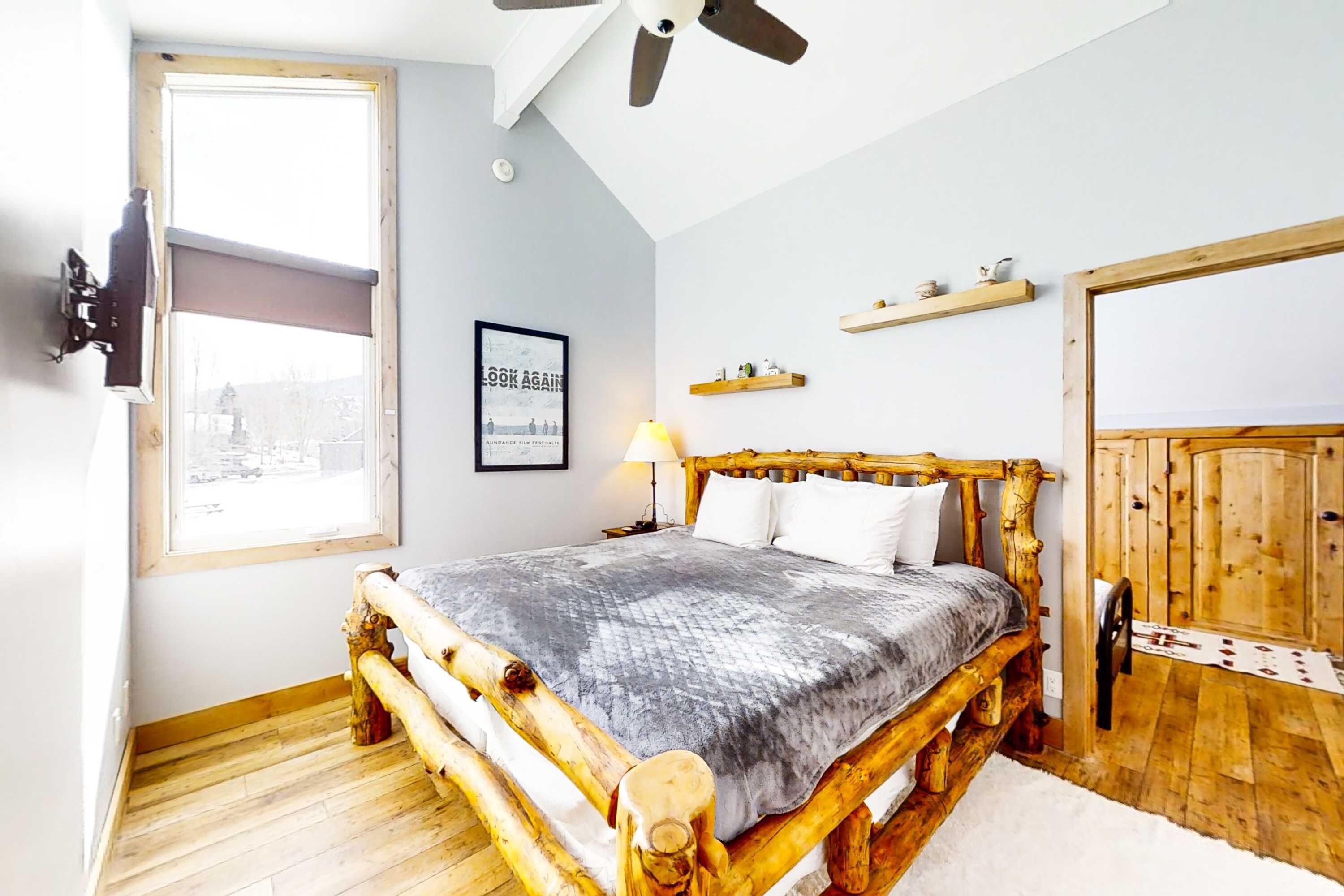 Ein Zimmer mit einem Bett, einem Deckenventilator, einem Fenster, einem gerahmten Bild und einer Holztür.