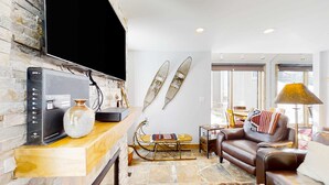 A living room with a stone wall, a flat-screen TV, a leather sofa, a coffee table, and a lamp. - Remodeled mountainside home with outdoor heated pool & hot tub - walk everywhere (Park City)