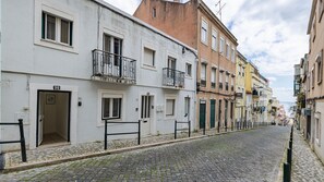 Eine Kopfsteinpflasterstraße gesäumt von mehrstöckigen Gebäuden, einige mit Balkonen und Fensterläden, und ein klarer Himmel darüber.
