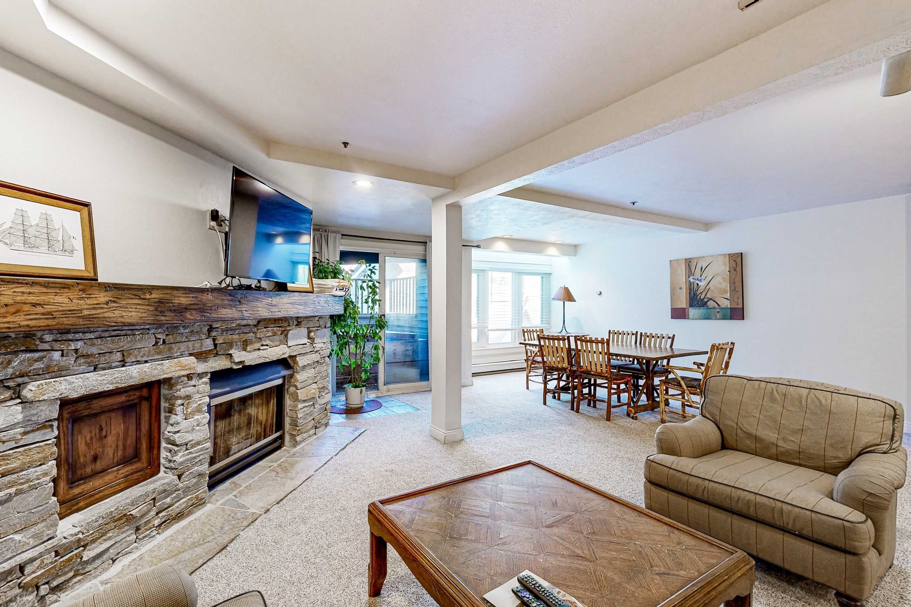 A living room with a stone fireplace, a wooden coffee table, a beige sofa, and a dining area with wooden chairs and a table.