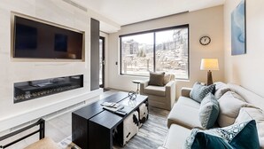 A modern living room with a fireplace, a sofa, a coffee table, and a large window offering a view of a mountainous area. - Pool Deck, Hot Tubs & Gym - Stunning Condo in New Ski-In, Ski-Out Community (Park City)