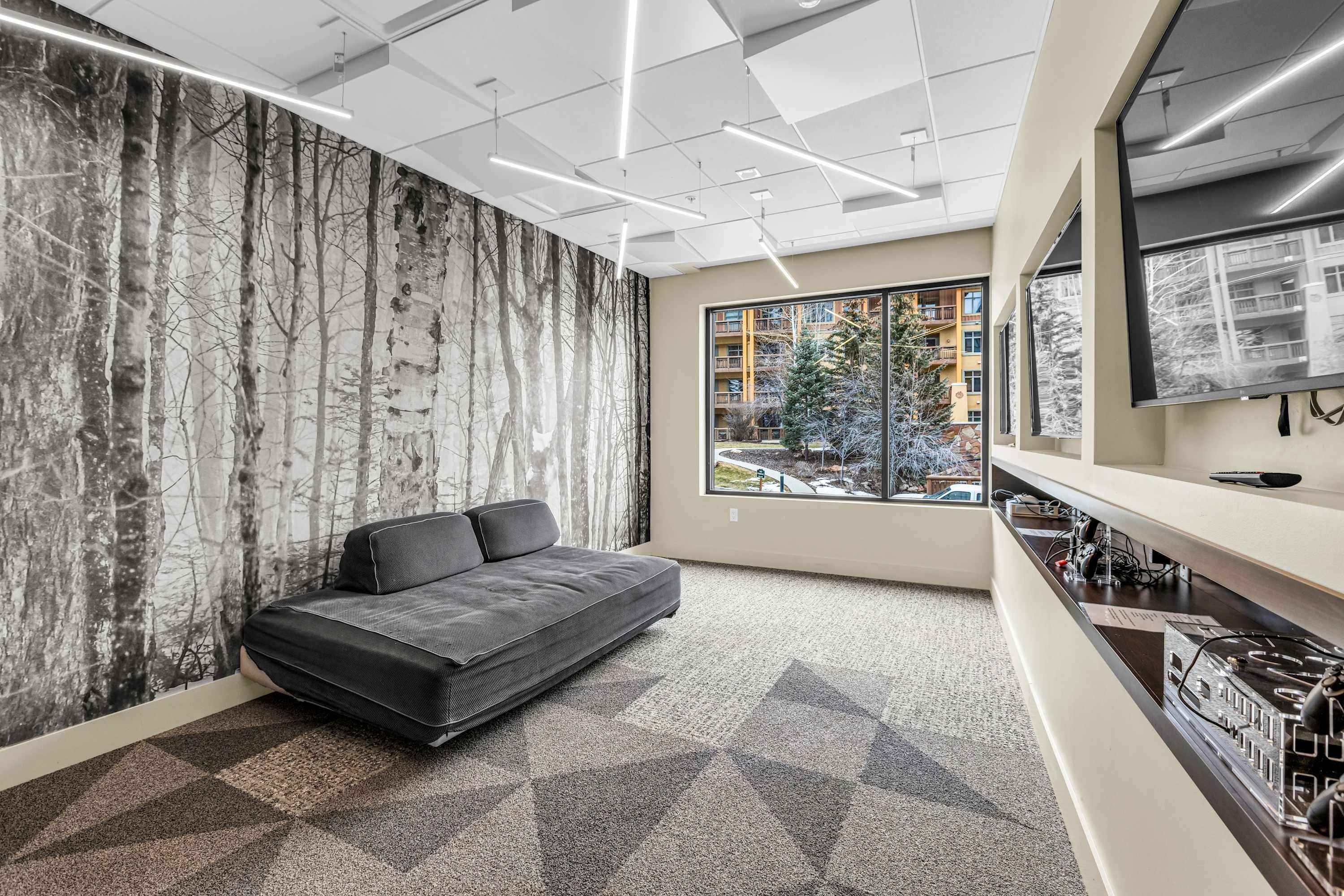 Un espace de bureau moderne, doté d’un canapé, d’une grande fenêtre offrant une vue sur les bâtiments et d’un mur orné d’une fresque représentant un arbre.