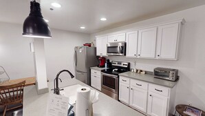 Een moderne keuken met witte kastjes, roestvrijstalen apparatuur, een zwarte hanglamp en een klein eethoekje met een houten tafel en stoel.