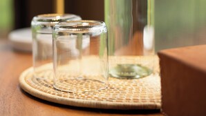 A clear glass bottle with a metal cap, two smaller glass containers, and a woven placemat on a wooden surface. - Mimark Serengeti Luxury Camp (Serengeti)
