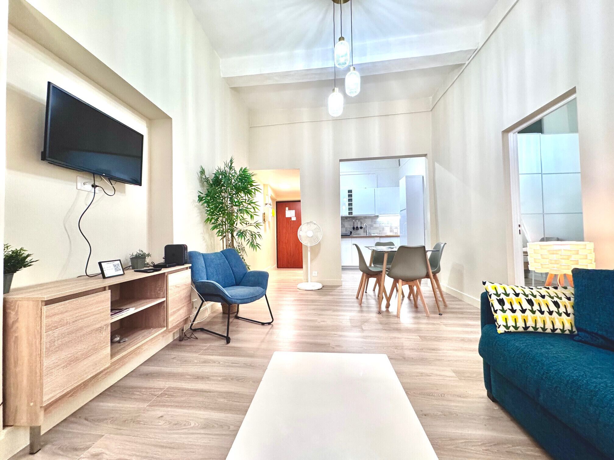 Ein modernes Wohnzimmer mit Flachbildfernseher, einem blauen Sessel, einer Essecke mit Tisch und Stühlen sowie einer Küche im Hintergrund.