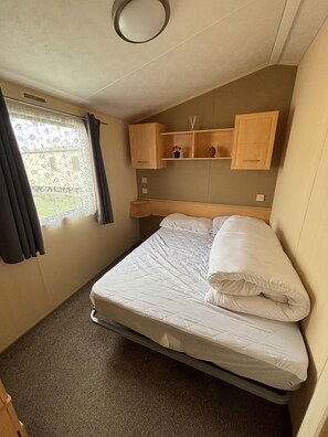 Una cama individual con colchón acolchado, una mesita de noche pequeña, una estantería con objetos decorativos y una ventana con cortinas.