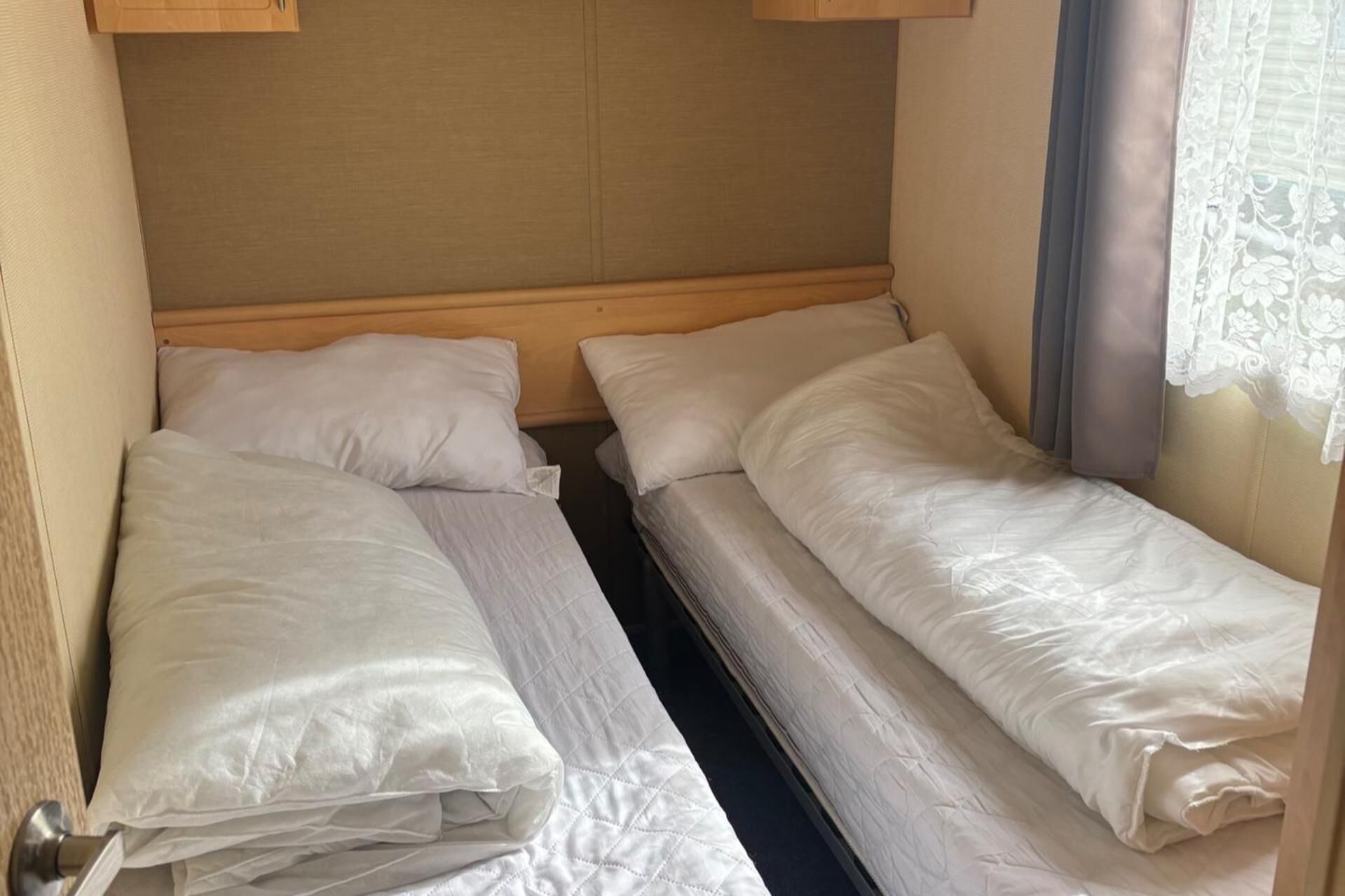 Una cama compacta con cabecera de madera y una ventana con cortinas de encaje.