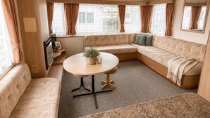 A cozy RV interior with a sectional sofa, a small table, and a window with curtains. - Seaside Tranquility Retreat in Clacton (Clacton-on-Sea)