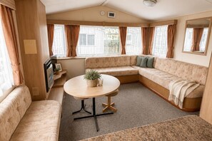 A cozy RV interior with a sectional sofa, a small table, and a window with curtains. - Seaside Tranquility Retreat in Clacton (Clacton-on-Sea)