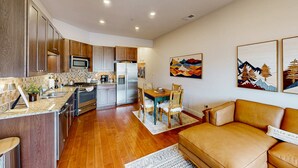 A modern kitchen with wooden cabinets, a microwave, and a refrigerator. There is a dining area with a table and chairs, and a sofa in the living room. - Inviting condo in Newpark Town Center with hot tub and great balcony views (Park City)