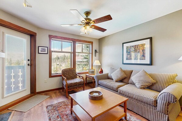 A living room with a ceiling fan, a patterned sofa, a wooden coffee table, a patterned armchair, and a window with a view of trees. - Mountainview Condo with Ski-In/Out Access, Club Solitude Access with Pool & Hot Tub (Solitude)