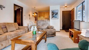 Een woonkamer met een bank, een bruine fauteuil, een salontafel, een tv-meubel met een televisie, en een keuken op de achtergrond.
