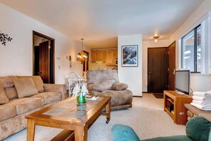 A living room with a sofa, a brown armchair, a coffee table, a TV stand with a TV, and a kitchen in the background. - Corner condo in Canyons - close to ski lifts w/ shared pools, hot tub, & sauna (Park City)
