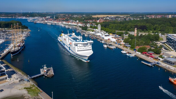 Ett stort kryssningsfartyg låg förtöjt vid en hamn med en småbåtshamn, byggnader och grönområden i bakgrunden.