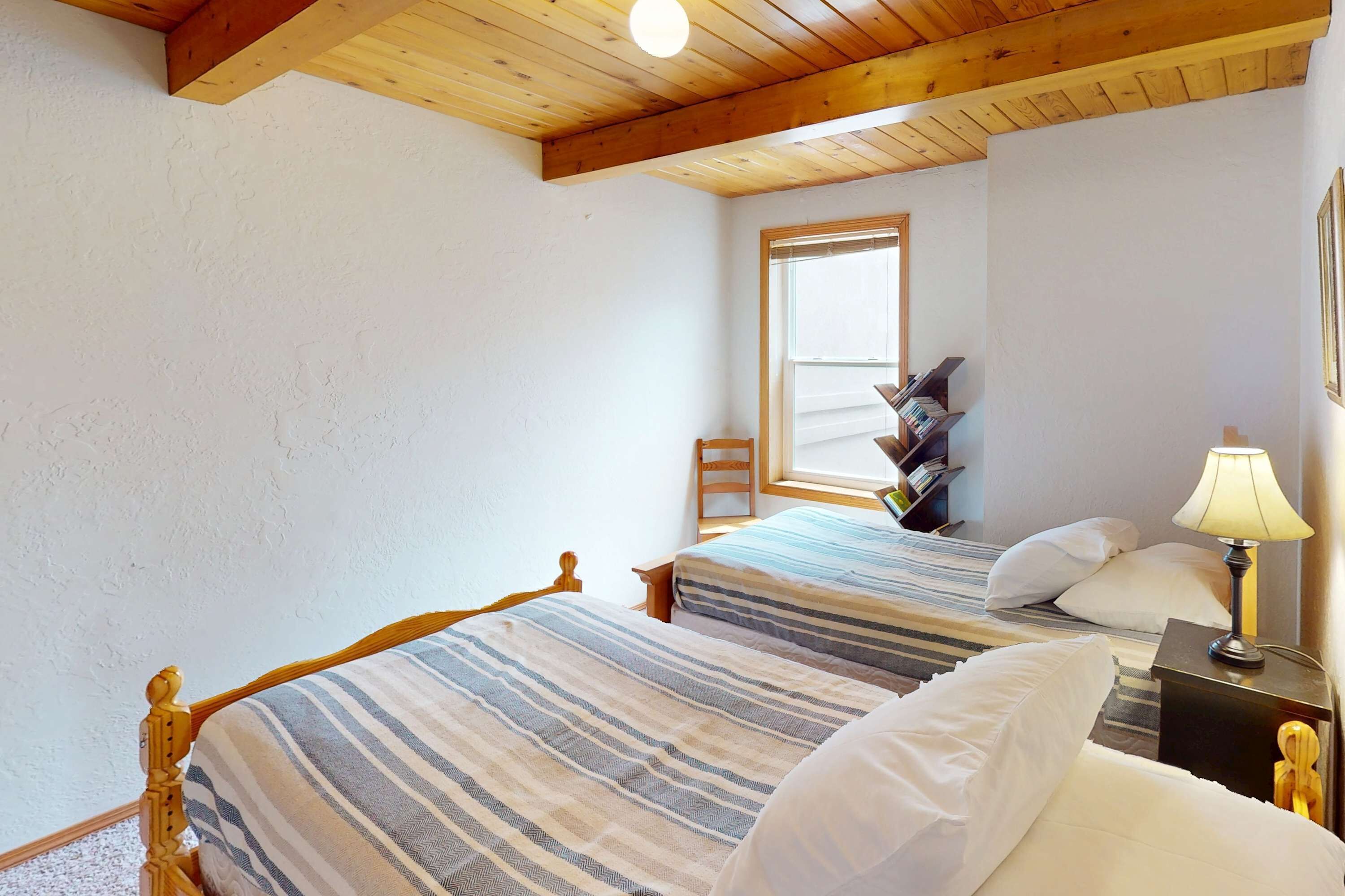 A bedroom with two beds, wooden headboard, a nightstand with a lamp, a window, and a chair.