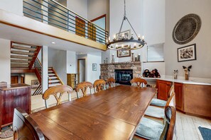 A spacious dining area with a long wooden table and chairs, a stone fireplace, and a staircase leading to an upper level. - Remodeled Bathrooms with ski theme, smart TVs, mtn views, grill, & hot tub (Park City)