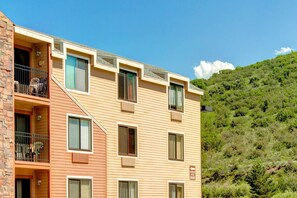 A multi-story building with balconies, a clear sky, and a hillside in the background. - Top-floor, mountain view condo with fireplace, pool & hot tub - steps to shuttle (Park City)