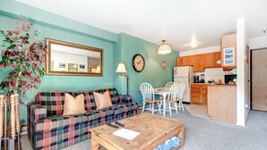 Ein Wohnzimmer mit karierter Couch, einem Couchtisch, einem Esstisch mit Stühlen, einer Küchenzeile und einem großen Spiegel an der Wand.