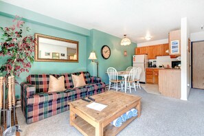 A living room with a plaid sofa, a coffee table, a dining table with chairs, a kitchen area, and a large mirror on the wall. - Top-floor, mountain view condo with fireplace, pool & hot tub - steps to shuttle (Park City)