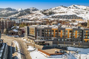 A hotel with multiple floors, a parking area, and a view of snow-covered mountains. - ski-in - ski-out condo in heart of Canyons Village with pool & hot tub access (Park City)