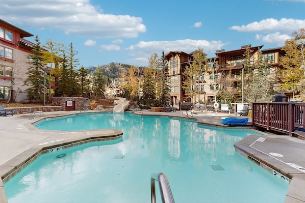 A swimming pool with a stone border, surrounded by buildings with balconies and a mountainous backdrop.
