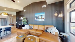 Un salon moderne avec un canapé d’angle, un coin repas doté de tabourets de bar, une cuisine équipée d’appareils électroménagers en acier inoxydable et une grande fenêtre laissant entrer la lumière naturelle.