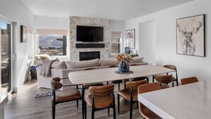 A modern living room with a stone fireplace, a flat-screen TV, and a dining area with a marble table and leather chairs. - Panoramic views, hot tub & game room, Minutes from Jordanelle Express Gondola (Heber City)