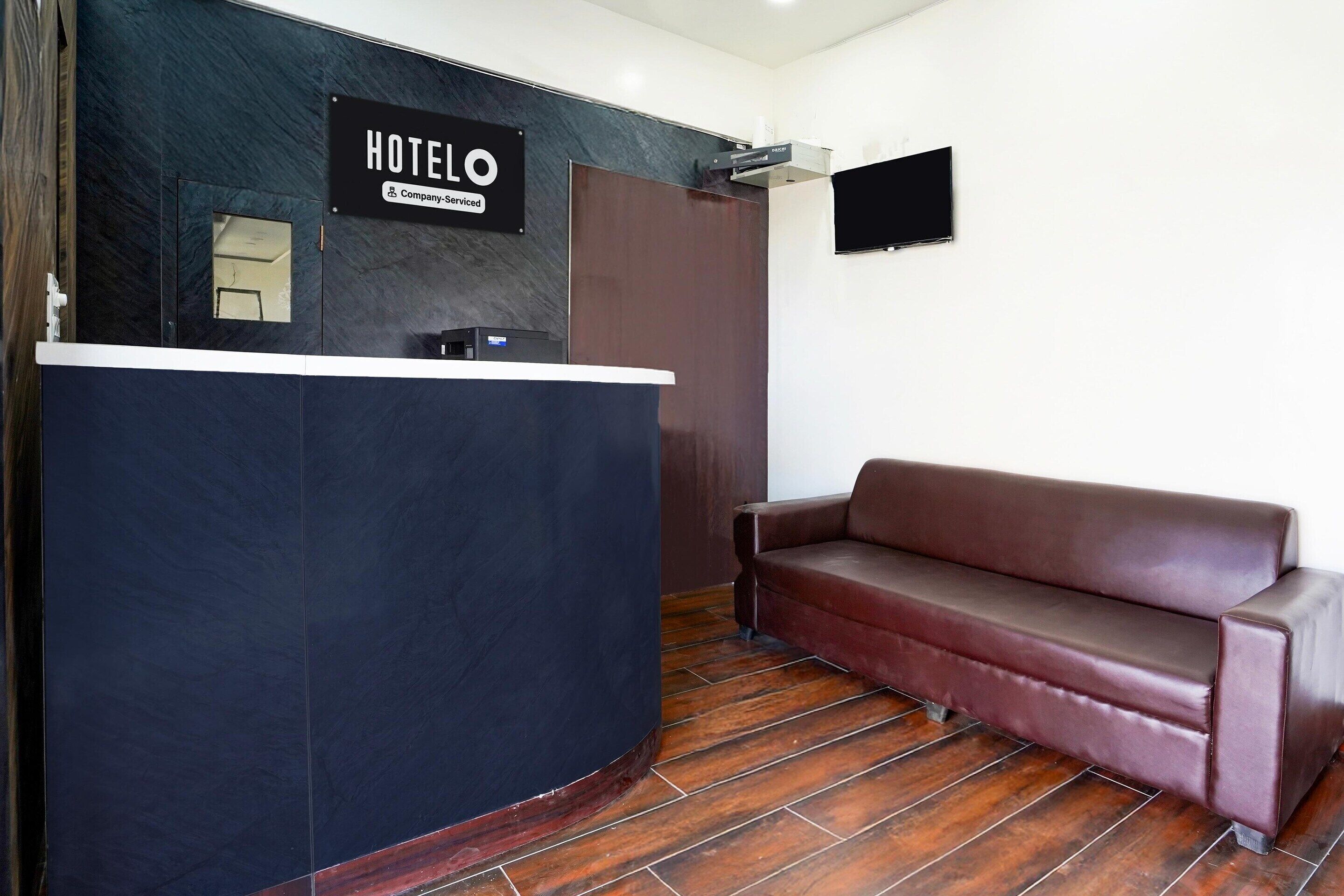 A hotel reception desk with a sign, a brown leather sofa, and a flat-screen TV on the wall.