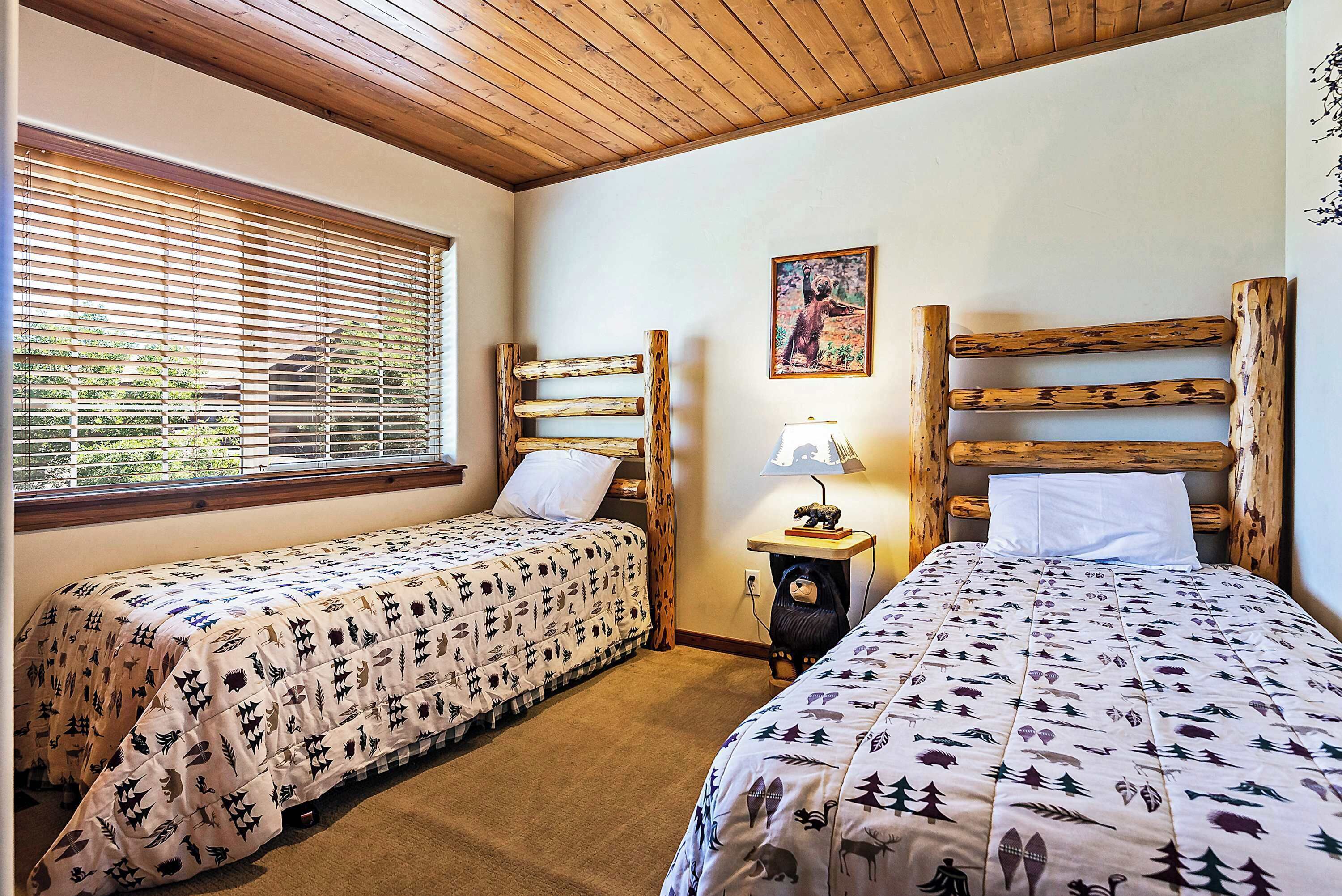 A room with two beds, wooden headboards, a window with blinds, a painting on the wall, and a lamp on a nightstand.