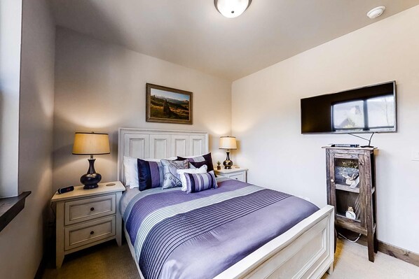 Un dormitorio con cama, dos mesitas de noche con lámparas, un televisor montado en la pared y un mueble auxiliar pequeño.