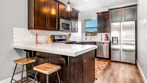 A kitchen with dark wood cabinets, stainless steel appliances, a white island with stools, and a view to the outside. - Private Rooftop Hot Tub Views Bumper Pool Table, High-Speed WiFi & Fireplace (Park City)