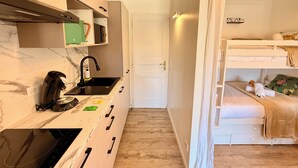 Eine kompakte Küche mit Kaffeemaschine, Mikrowelle und Spüle. Eine weiße Tür führt zu einem Schlafzimmer mit Etagenbett.